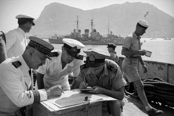 Archival black-and-white silver-gelatin press photograph taken in summer 1940 aboard a French cruiser in Gibraltar harbor; shot on a Leica IIIa with a 50mm uncoated Summar lens on Kodak Super-XX film; noticeable medium grain, slight halation around sunlit edges, mild edge vignetting, hand-held at roughly 1/125; asymmetric, candid composition with a knot of distinct French and British naval officers in summer whites and khaki bent over a small chart on a scrubbed quarterdeck; one older French captain with a trimmed moustache points with a pencil, a younger British lieutenant jots notes on a clipboard, a petty officer passes with a dented enamel mug; background shows the Rock of Gibraltar rising hazy, two Royal Navy destroyers at anchor with gulls wheeling, coiled rope, chipped paint on railings, light surf glitter; no signage or legible text anywhere in frame.