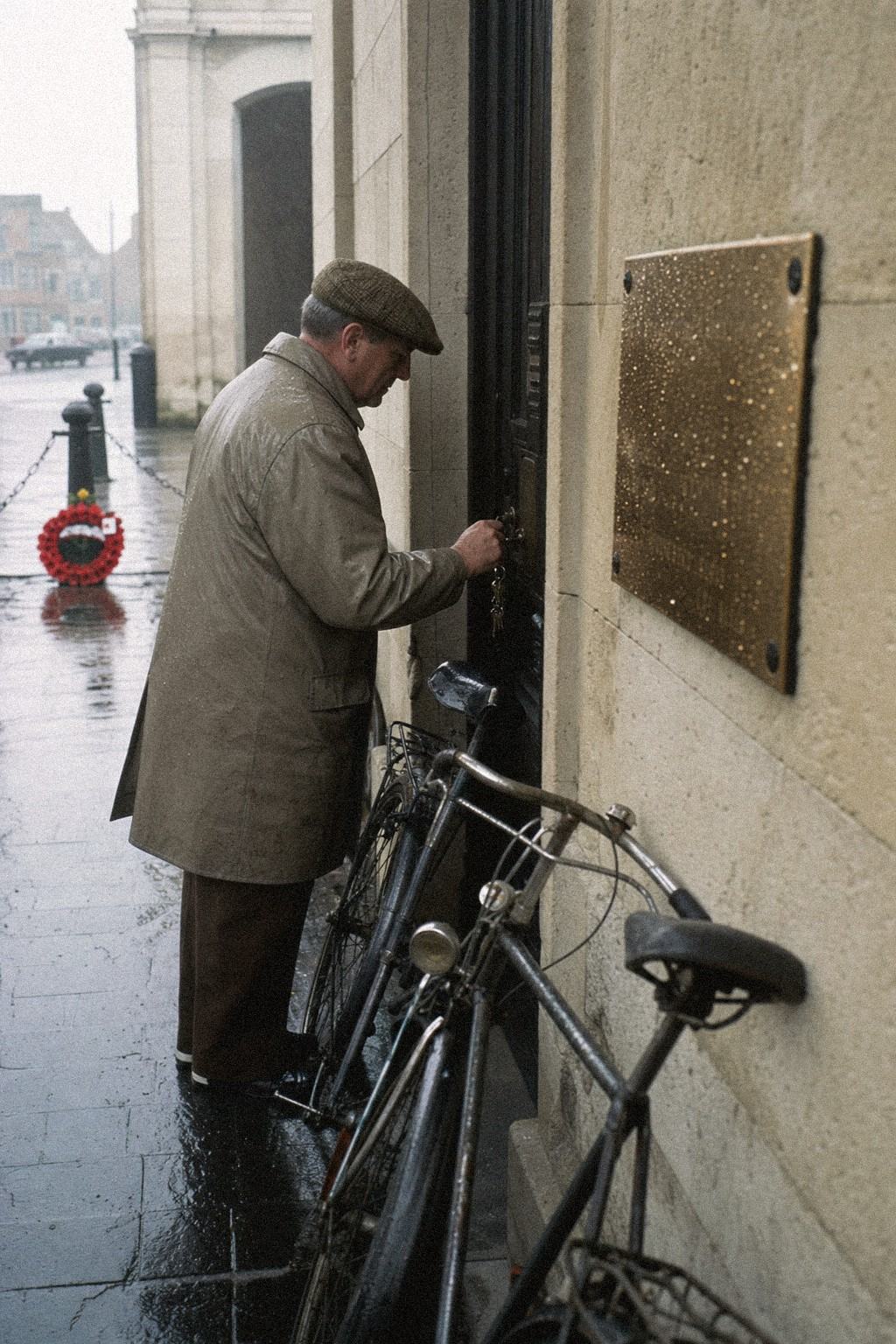 At Menin Gate House, a clerk unlocks a side door as bicycles lean against wet limestone in light rain.
