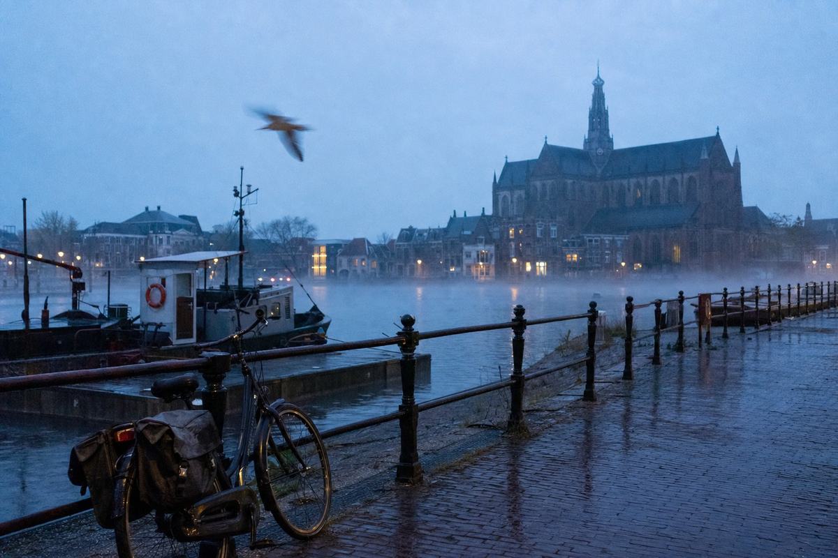 Dageraad aan de Spaarne met de Grote of Sint‑Bavokerk; natte kasseien, een afgemeerd bootje en lichte nevel boven het water.