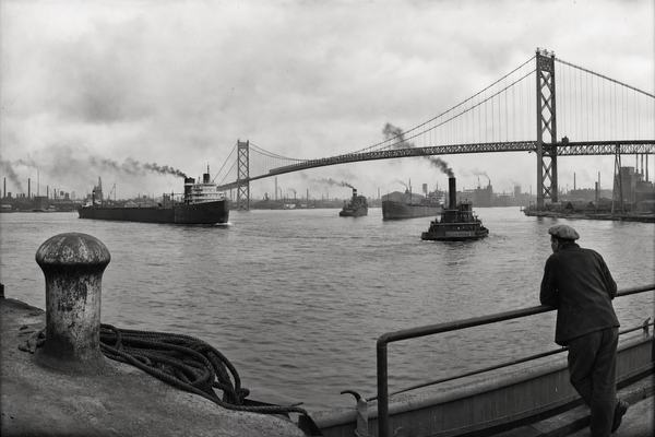 Archival silver gelatin panoramic photograph shot in the early 1930s using a Cirkut No. 10 rotating panoramic camera with an 8-inch lens; fine-to-medium film grain, slight edge vignetting, gentle barrel distortion across the sweep. Asymmetric composition from the Detroit shoreline with a weathered mooring bollard and coiled rope in the near left foreground; the Ambassador Bridge arcs from the right third toward the left distance. Multiple lake freighters and a tug move through the Detroit River channel, soft coal smoke drifting low. Assembly plants, railyards, and smokestacks line both the Detroit and Windsor, Michigan banks under overcast light. One dockworker in a flat cap leans on a railing, turned three-quarters away. Tonal range balanced for sky and water; no legible signage anywhere, realistic documentary style.
