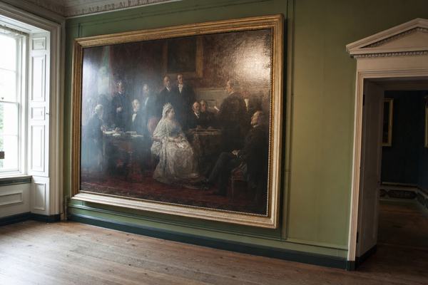 Contemporary digital press photograph inside a quiet Windsor Castle gallery in 2011, taken with a full-frame DSLR (35mm lens at f/4). Off-center composition places a large Victorian oil painting of Queen Victoria with her 1861 cabinet slightly left of frame, in a gilded frame. Soft natural side light from a tall window creates gentle reflections on varnish and subtle glare on the canvas; muted green walls and polished wooden floorboards show scuffs and fine dust. No people or signage are visible. Slight high-ISO noise in the shadows, mild barrel distortion, and shallow depth of field keep background molding soft while the painting remains crisp. The room’s doorway on the right edge reveals a darker antechamber.