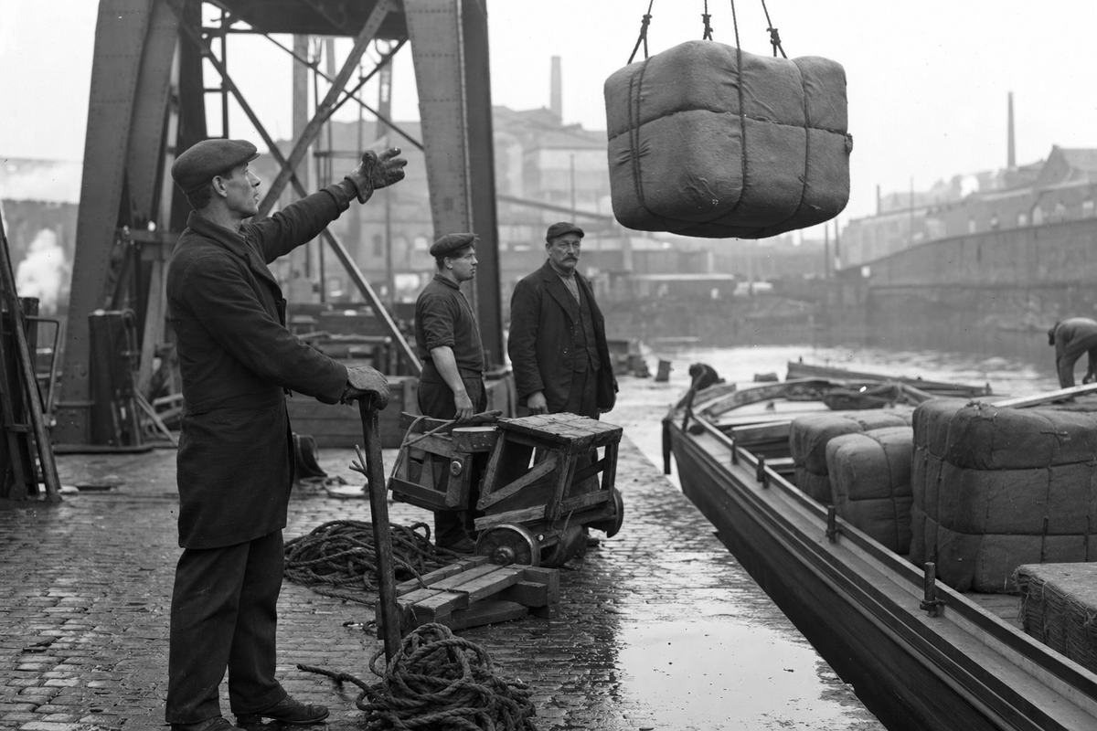 Early 20th-century dockside photograph in Manchester, c. 1905, silver gelatin print from a glass plate negative with moderate contrast and soot haze softening distant warehouses. Asymmetric frame shows a stevedore at left foreground—lean, mid-40s, flat cap—gesturing upward with a gloved hand as a single cotton bale is hoisted by a crane; rope coils and wooden pallets spill across wet cobbles. Two other workers—one younger, broad-shouldered, another older with a moustache—wait near a handcart; faces and builds are distinct. A low canal barge sits to the right edge with stacked, unmarked bales turned so no stenciling is visible. Puddles reflect gantry beams; no text or signage appears.
