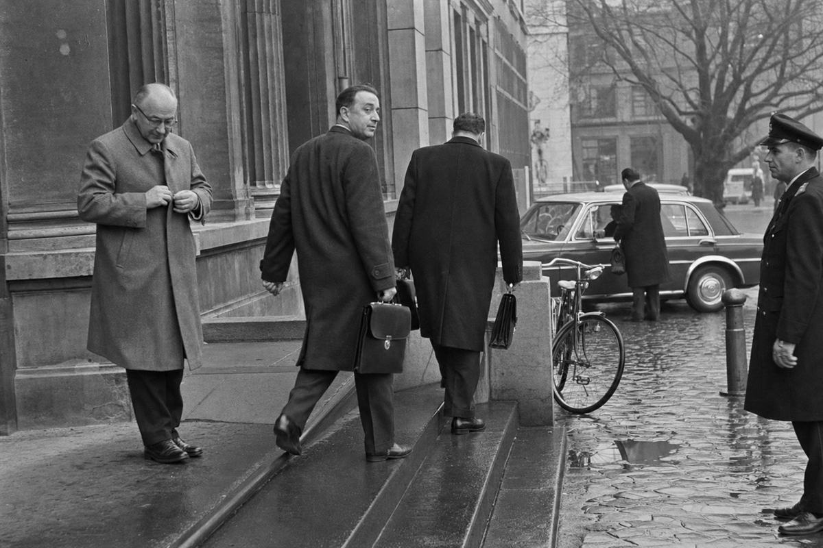 1960s black-and-white press photograph on Kodak Tri-X 400, shot with a 35mm Leica M3 and 35mm lens, capturing members arriving at the Federal German Union’s parliament in Frankfurt. Asymmetric street-level angle from the foot of the steps; two suited deputies in overcoats stride up with briefcases, one glancing back while a third, older member pauses to button his coat. Stone facade and columns frame the scene; wet cobbles show shallow puddles, a bicycle leans against a low railing, a taxi idles beyond with number plate blurred, a uniformed constable at the edge. Moderate grain, slight motion blur in legs, crisp highlights on wet stone; no visible signage or text.