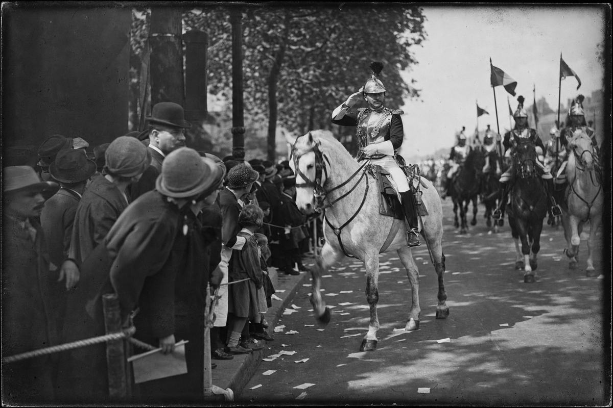 Silver gelatin print from a 9x12 cm glass plate negative, handheld folding plate camera with ~135mm lens, Paris circa 1930; curbside oblique view of a Waterloo Day parade on the Champs-Élysées: a cuirassier officer on a light-gray horse turns to salute, motion blur at the hooves; spectators in hats lean forward behind a rope, scattered paper on the pavement, tree shadows crossing the scene; fine grain with moderate contrast, slight edge vignetting and minor emulsion scratches; uniforms accurate, flags without any text; no signage or lettering visible.