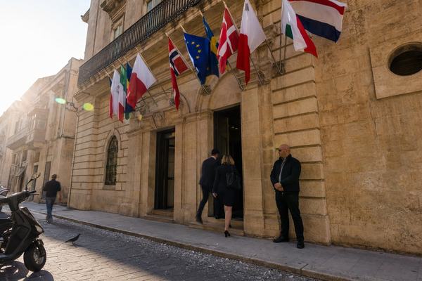 Digital press photograph captured in early morning light in June 2026 on St. Christopher Street, Valletta; full‑frame DSLR with a 35mm lens, handheld at 1/250s, ISO 200. Oblique low angle from across the narrow limestone street showing the Mediterranean Patrol Authority headquarters facade with multiple member flags fluttering. Two staff members in business attire step through the doorway while a plain‑clothes security guard watches the street; slight motion blur on the flags and a passerby. Warm limestone tones, mild lens flare at frame edge, pavement scuffs, a partly cropped parked scooter, and scattered pigeon feathers on the curb. Exposure balanced for highlights; no text or signage visible.