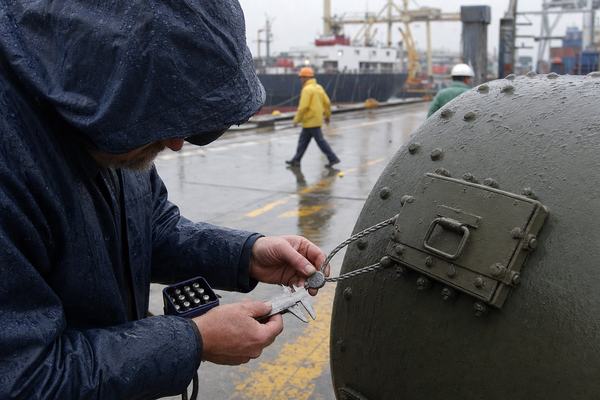 Digital press photograph captured in 2003 with an early-2000s DSLR (high ISO noise, slight motion blur in rain), 50mm lens at f/2.8, natural overcast light. A marine insurer’s surveyor in dark rain gear leans over the right flank of a Type B(U) transport cask on a wet Pacific port quay, using metal calipers and a small steel stamp set to verify and mark a numbered lead seal crimped on a braided stainless cord. Asymmetric composition places the surveyor at the left third, the slick curved cask dominating the right; visible water droplets bead on painted metal, rain ripples in shallow puddles. Yellow deck markings and rust stains cut across the concrete; a moored cargo vessel and container cranes sit softly out of focus in the background, with a couple of dockworkers blurred mid-stride farther back. No legible text or signage; the scene feels candid and worklike.