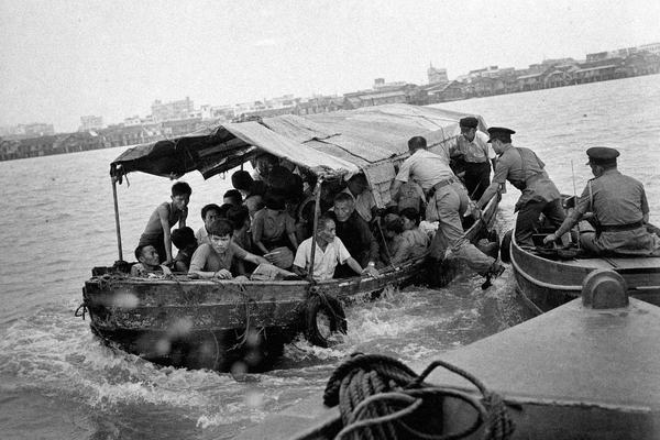 Black-and-white 1930s press photograph taken at first light from a low angle on a small boat, showing an overloaded, low-freeboard motorized tongkang mid-channel between Butterworth and George Town. Two Marine Police launches are tight alongside as officers board; passengers of varied ages and builds huddle beneath a patched canvas awning, a few men gripping the gunwale with strained postures and sweat-sheened faces. Early tide chop flicks spray against the lens, producing slight flare and halation; horizon subtly canted. George Town’s skyline and clan jetties recede in humid haze; floating debris and a coiled rope in the foreground add depth. Silver gelatin press print from a 4x5 panchromatic sheet negative, shot on a Graflex Speed Graphic with a 127mm lens; moderate grain, slight motion blur on hands and water, uneven exposure at the frame edges. No modern elements, no visible text or lettering, no watermarks.