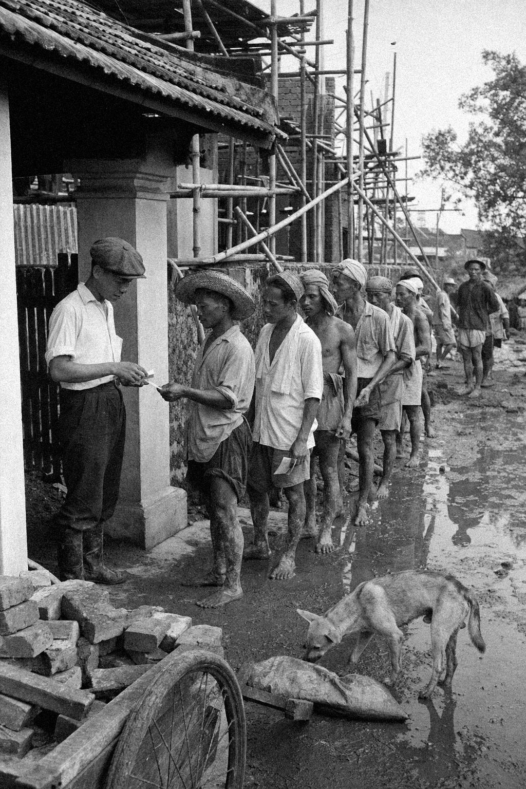 At Sia Boey, labourers queue at a side gate with paper passes while a foreman checks them beneath bamboo scaffolding.