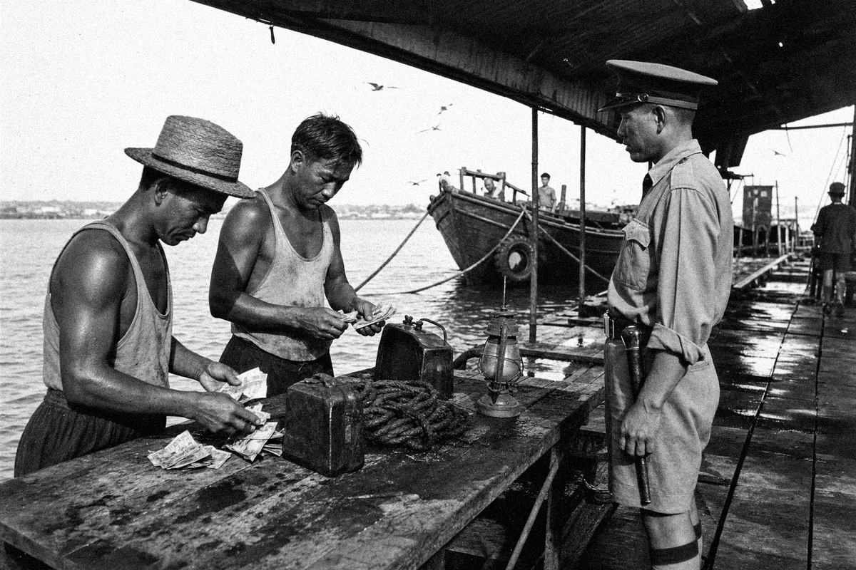 At Weld Quay, lightermen count cash beside jerrycans and rope while a constable watches from the shade.