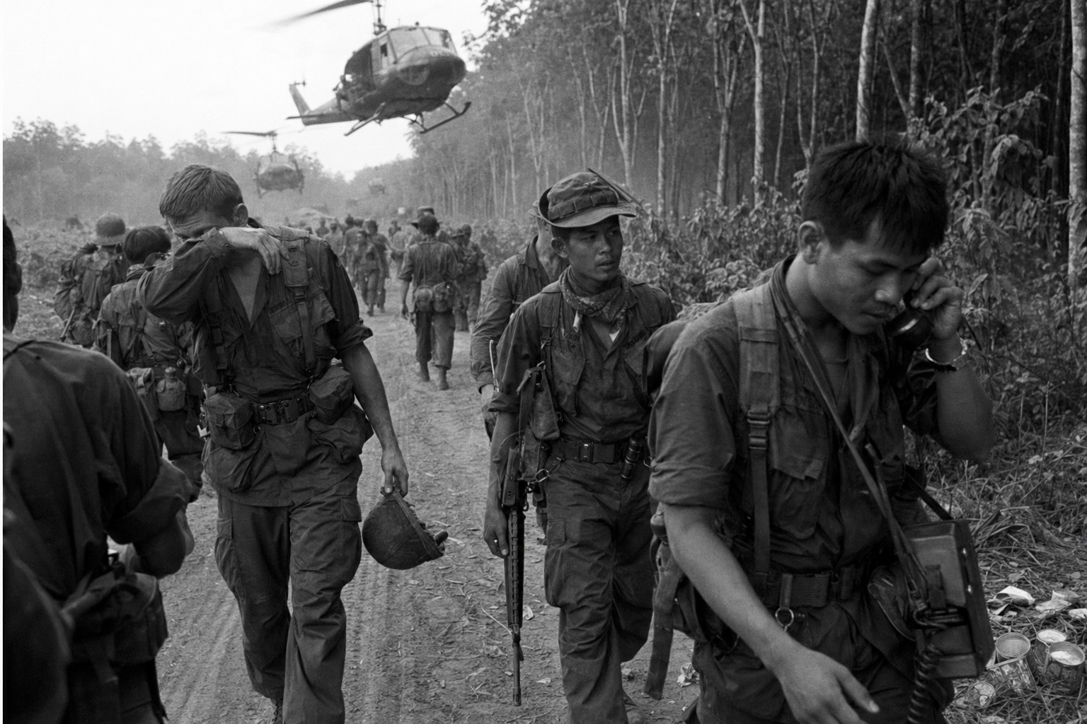 CMFF troops from U.S., ARVN, and Thailand advance on a red-dirt road near Chup as helicopters throw dust into the rubber trees, 1971.