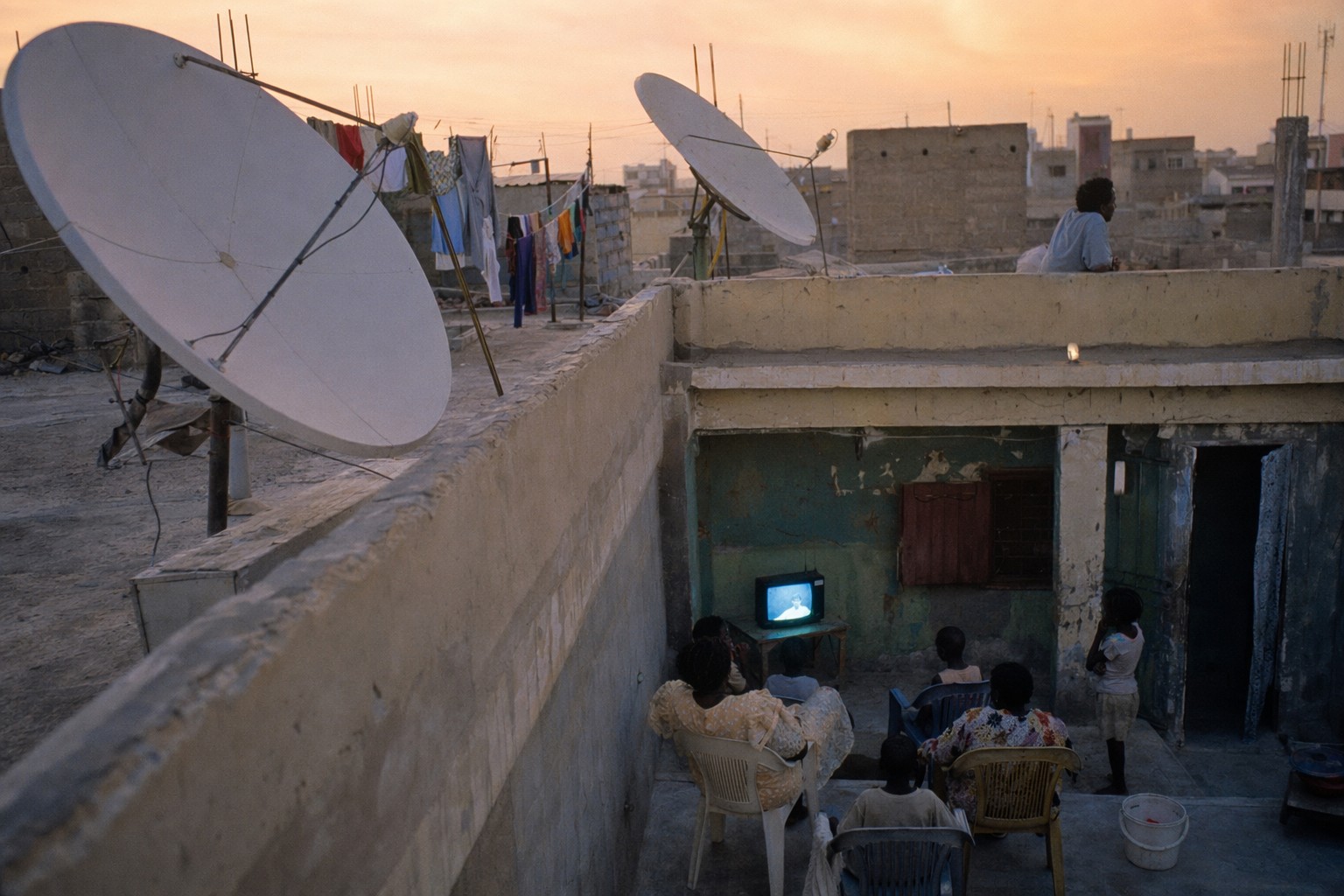 Toits de Dakar avec paraboles orientées vers la ceinture géostationnaire et famille regardant la télévision dans la cour au crépuscule.