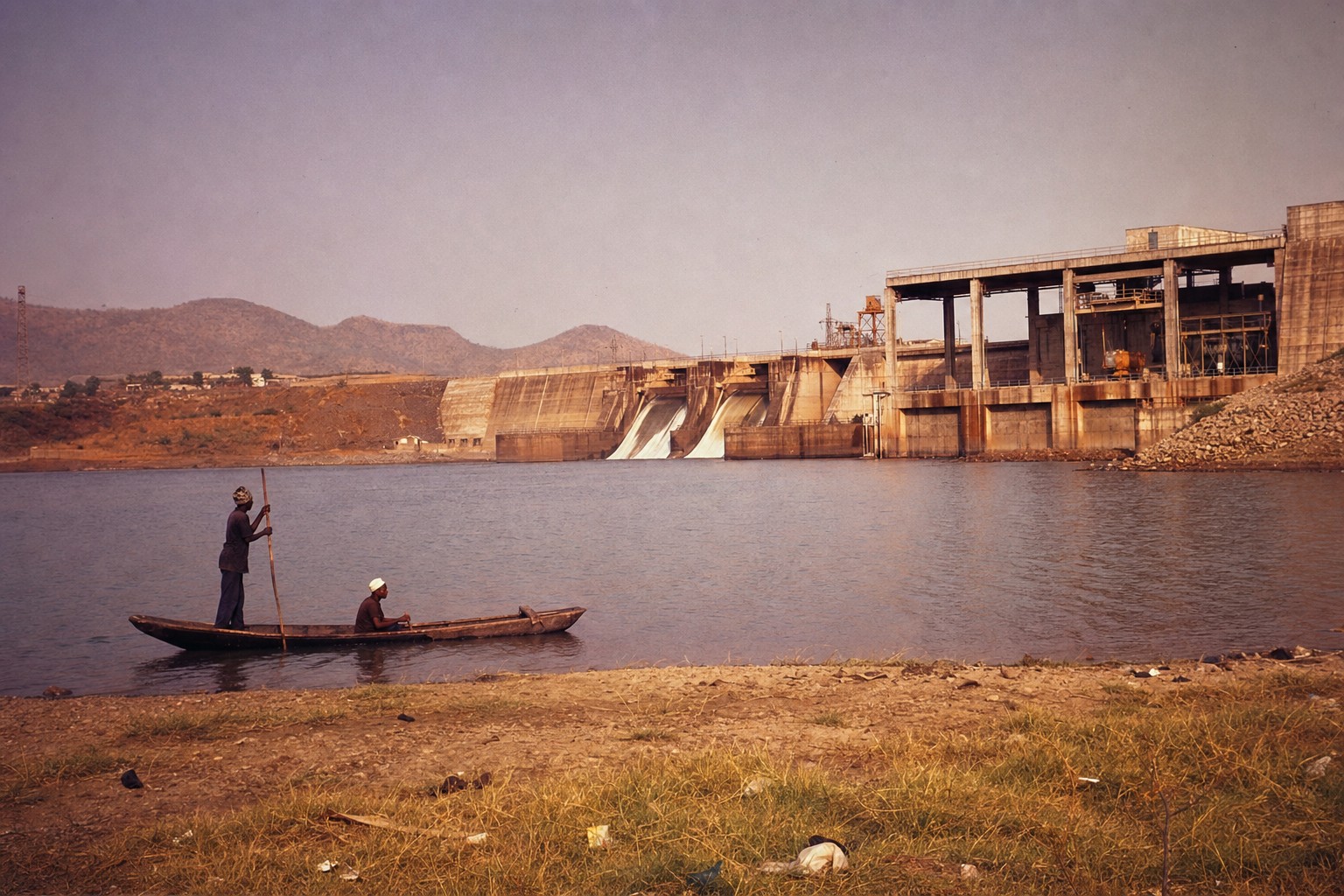 Vue oblique du barrage de Manantali à la fin des années 1970, avec une pirogue au premier plan et la centrale en arrière-plan.