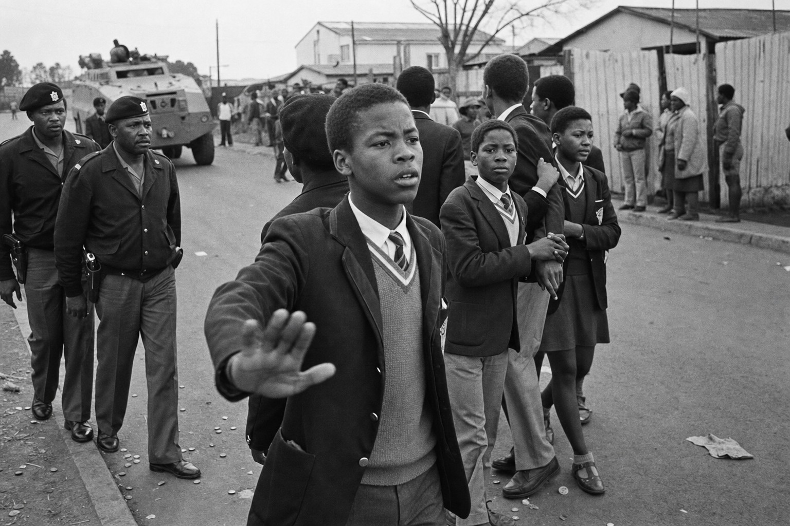 Students march on Vilakazi Street under police escort as a student marshal signals to keep formation; a Saracen vehicle holds back at an intersection.